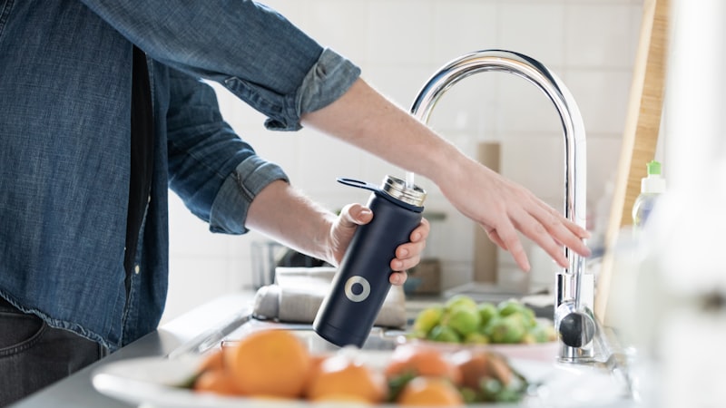 Person refilling a reusable water bottle at a kitchen sink, the refill a hydration tracker counts
