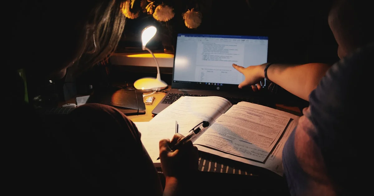 Student using a note-taking app on a laptop beside open textbooks at a desk lamp