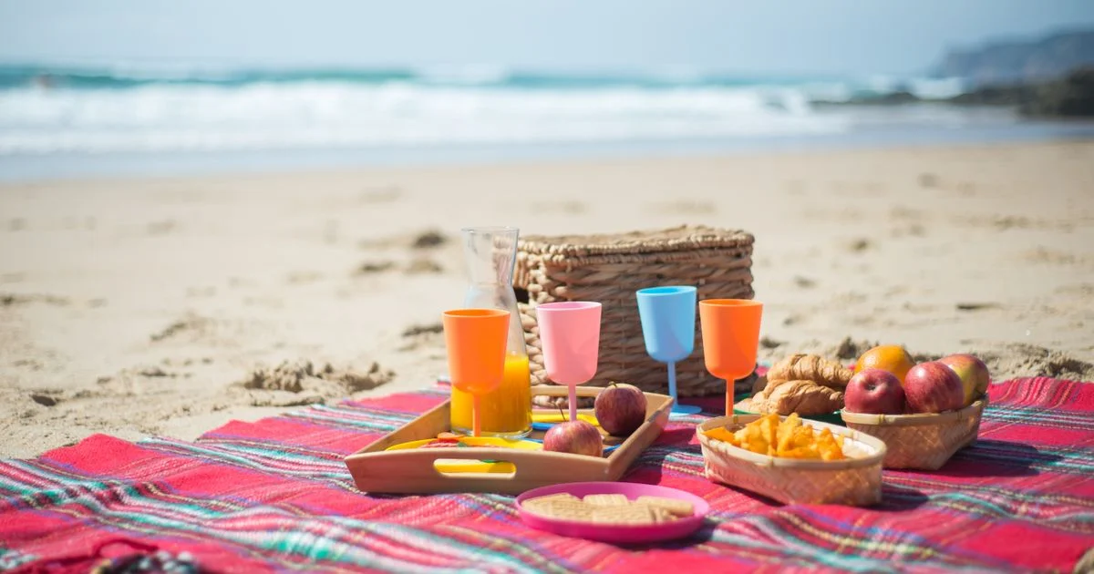 A beach picnic laid out on a blanket on open sand beside the surf, the sandy setting a bluetooth speaker for outdoor use has to survive