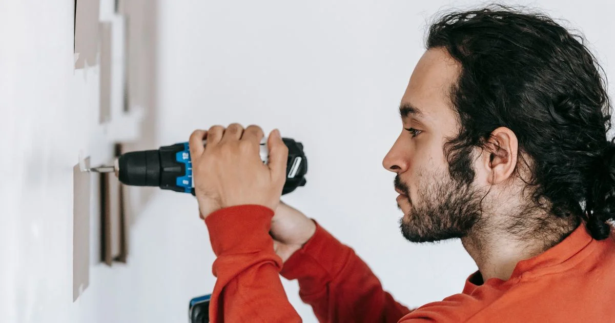 A man driving a screw into a marked interior wall to mount a board, the first real job for a cordless power drill for beginners
