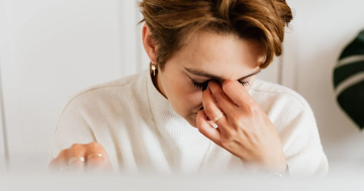 A woman at a bright desk pressing the skin beneath her eye, the tired-at-a-screen moment that sends people looking for an eye massager for dark circles