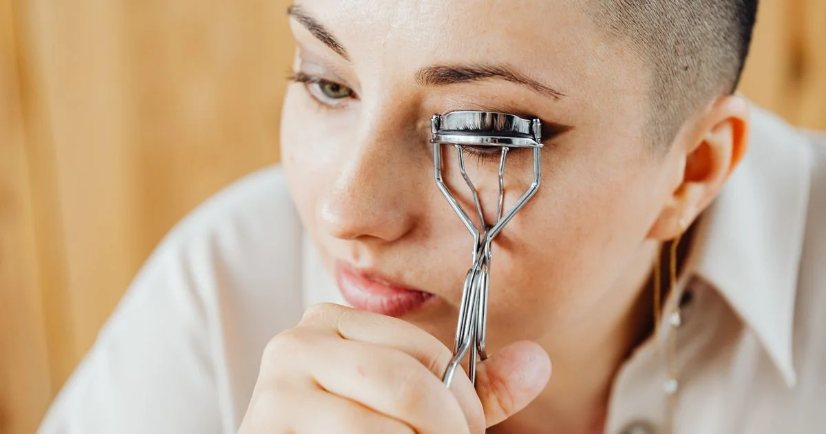 A woman clamping a metal manual curler on her upper lashes in daylight, the daily step a heated eyelash curler for straight lashes is meant to shorten