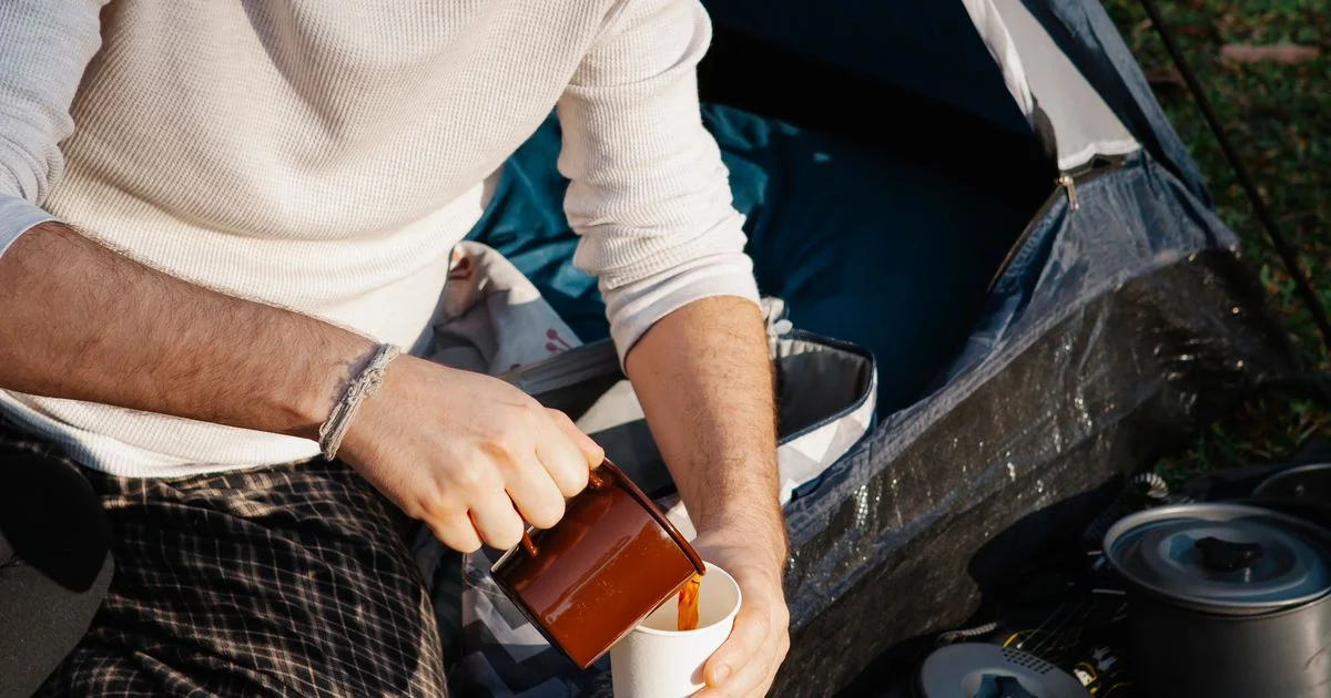A camper pours coffee into a cup beside a tent, the situation a portable espresso maker for travel is bought for