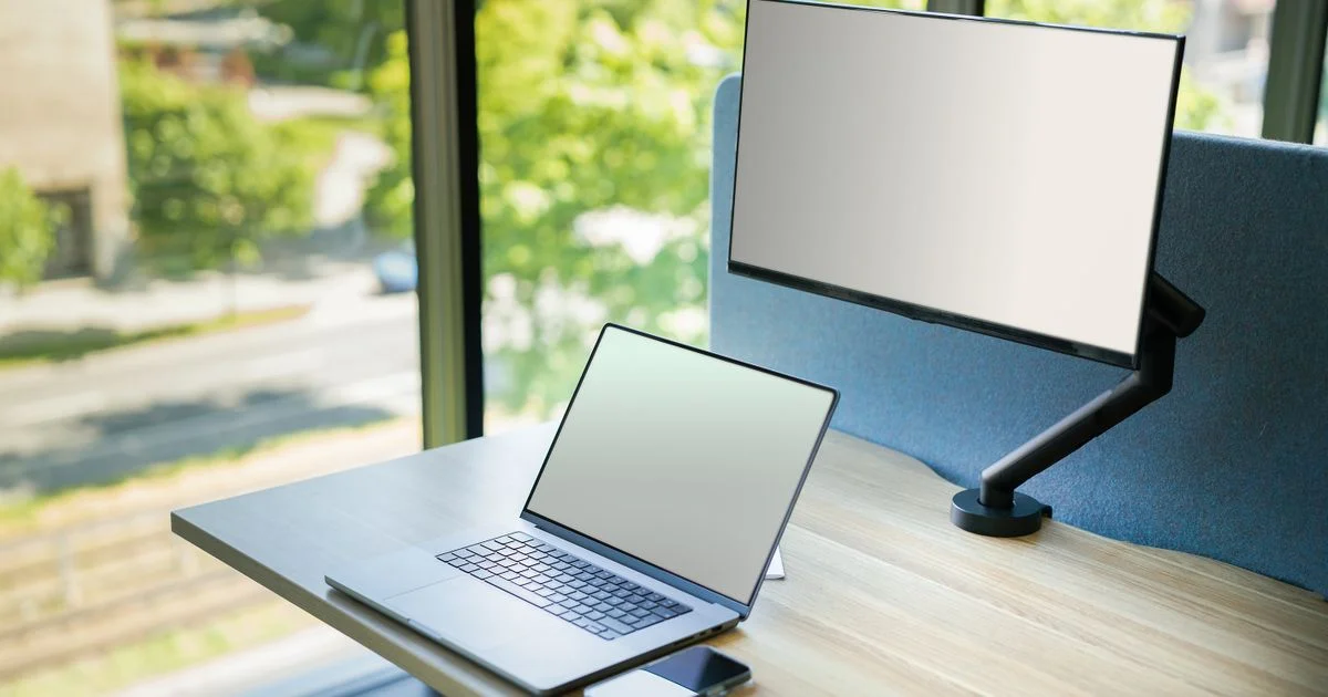 A laptop beside a slim second screen on a desk by a window, the setup a portable monitor for laptop work replaces