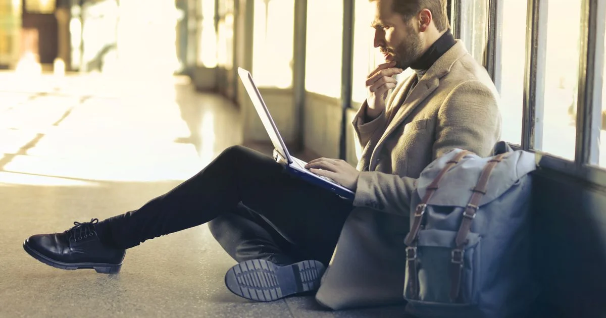 Traveler working on a laptop on the floor beside his bag at an airport window, a smart backpack for travel in everyday use