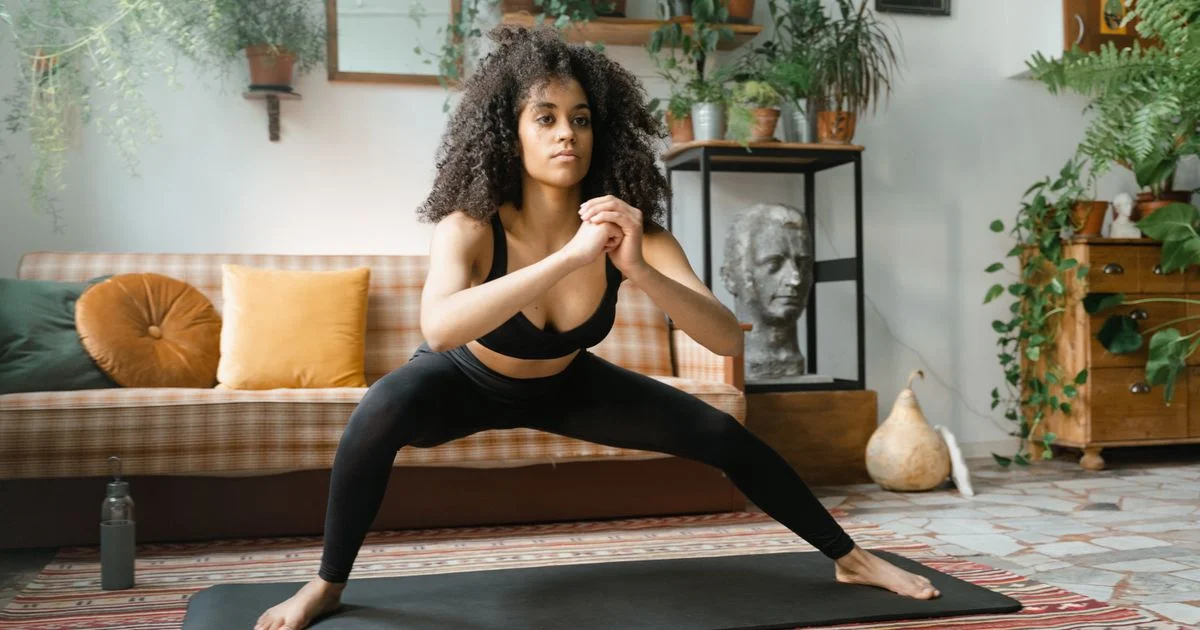A woman holding a wide-legged standing pose on a plain mat in a crowded living room, the home setting a smart yoga mat for beginners is bought for