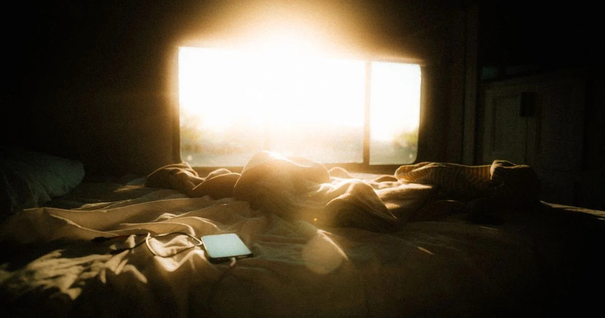 Early sunlight filling a dark bedroom over an unmade bed, the effect a sunrise alarm clock for waking up naturally tries to copy